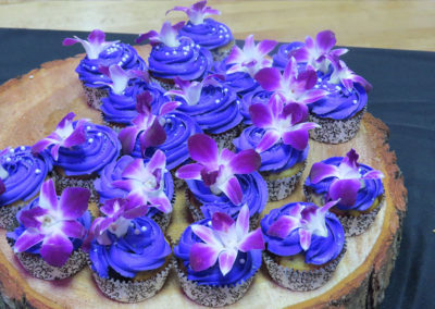 Blueberry cupcakes with purple frosting, garnished with a purple flower. Served on a rustic wood board.